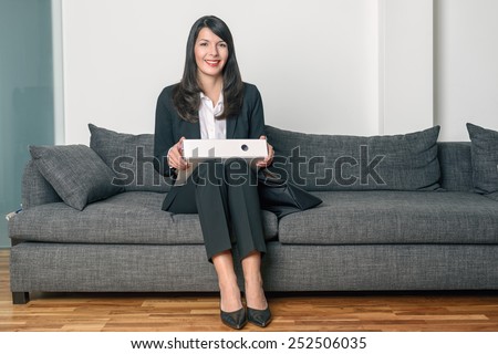 Smiling stylish businesswoman sitting on a grey sofa holding a binder across her knees as she waits for an appointment or meeting, with copy space