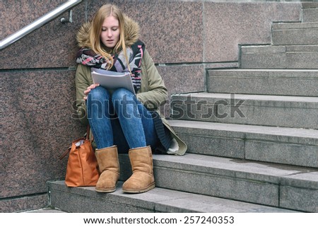 Trendy young woman in winter fashion sitting reading on outdoor, urban steps as she studies up her college notes or reads a journal while waiting for someone