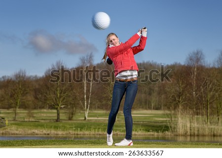 Woman golfer hitting the golf ball with a driver in front of a water hazard on a golf course with the ball flying through the air towards the camera