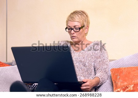 Serious Middle Age Woman with Short Blond Hair Sitting on Sofa While Using her Laptop Computer.