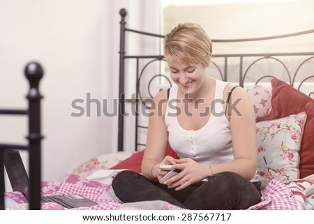 Young pretty blond teenage girl sitting cross-legged on top of her wrought iron bed texting on her mobile phone smiling with pleasure