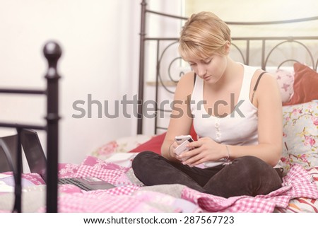 Young pretty blond teenage girl sitting cross-legged on top of her wrought iron bed texting on her mobile phone with a serious expression