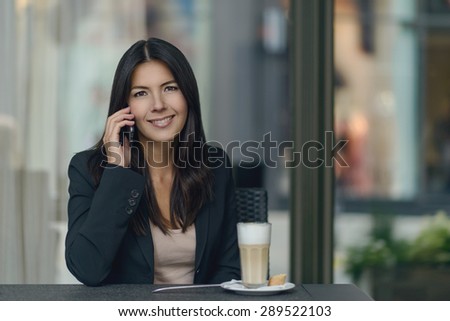Smiling woman chatting on her mobile phone while sitting in a cafeteria enjoying a delicious cup of cappuccino coffee giving the camera a friendly smile