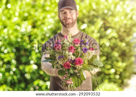 Three quarter length front view of smiling bearded 20s man wearing brown baseball cap, brown t-shirt and jeans holding bunch of flowers.