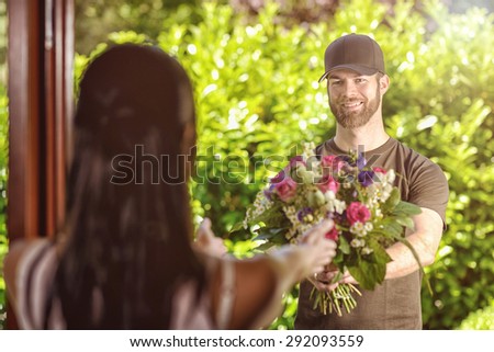 Smiling bearded 20s man wearing brown cap and brown t-shirt delivers flowers to door of young brunette female. Over the shoulder rear view of brunette.