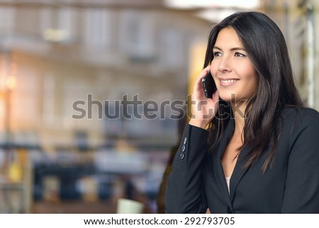Smiling Young Businesswoman in Black Suit Listening to Someone on Mobile Phone, Beside a Glass Window of a Shop, While Looking Into Distance.
