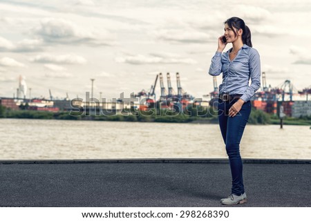 Attractive brunette happy woman wearing casual outfit as blue skinny jeans and shirt, while talking on mobile phone and walking on shore along the river in a cloudy day