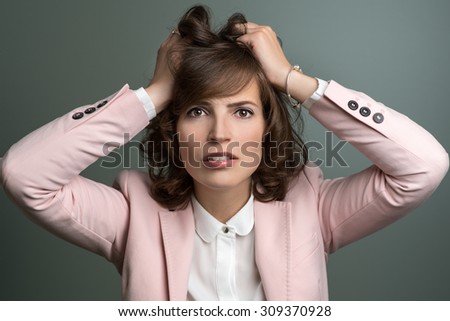 Attractive stylish young woman in a pink jacket tearing at her brown hair with her hands as she vents her frustration, over grey