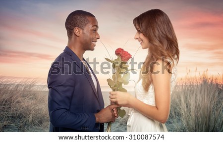 Smiling romantic handsome young African man proposing to his sweetheart as he clutches a bunch of long stemmed red roses in his hand, sideview in the sunset on a beach