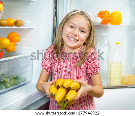 Smiling little girl puts bananas in the refrigerator with fresh vegetables