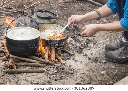 tourist man fry food in a pan over campfire in the forest during hike, cauldron hanging beside, horizontal version.