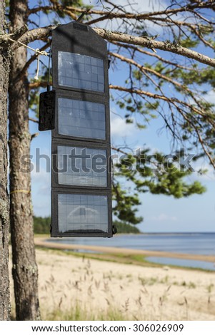 Cell mobile phone charges via USB cable from the portable foldable solar panel battery hanging on the outdoors on a pine tree on the seashore near the sandy beach at camping