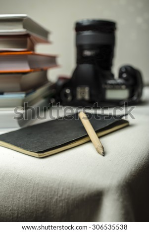 modern digital SLR camera on the table next to a stack of books, and in the foreground a notebook and pencil