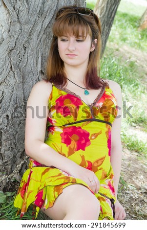 Beautiful girl in a dress sitting and leaning against a tree looking to the side and down