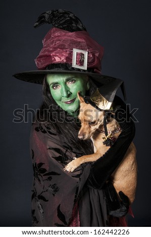 Halloween witch with green face holding small dog wearing wizard hat on red background
