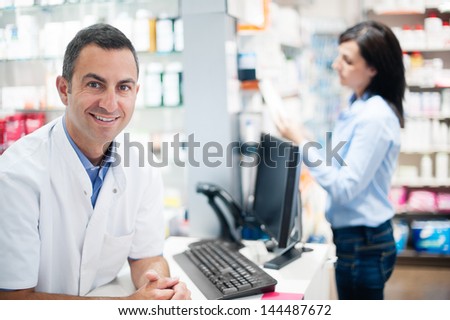 In the foreground, a pharmacist looks pleased to camera. In the background, a customer is inquiring about medication