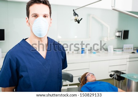Dentist in his surgery. In the background we can see the patient. He poses looking at camera