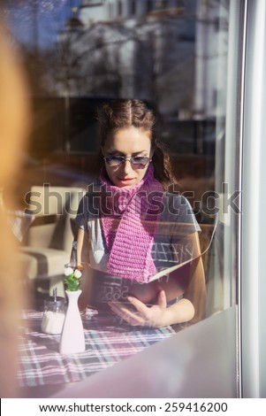 A beautiful confident young woman with stylish sunglasses, sitting in restaurant. Shoot trough the window/Young adult woman in restaurant