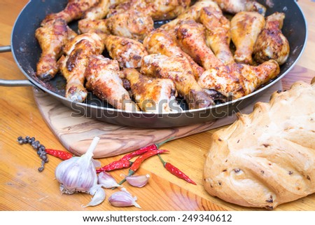 Roasted chicken drumsticks on a frying pan on the cutting board, with garlic and chilly pepper and bread around on a wooden table