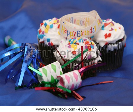 three cupcakes with happy birthday ribbon and noise makers on blue satin fabric with selective focus on front cupcake and ribbon