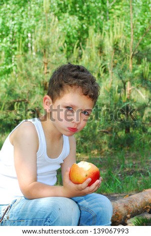 In the summer, bright sunny day, the boy sits in a pine forest on a log and eats an apple