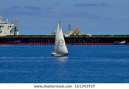 Small sailboat sailing on the bay and next to large supertanker docked