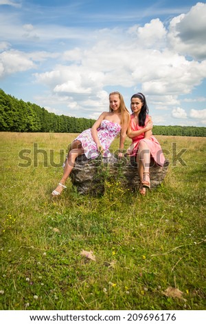 Two beautiful girls in the field. Girl sitting on a rock. They smile, radiate positive energy here, communicate. nature, beauty, girl, girlfriends - summer vacation concept. Article about the weekend.