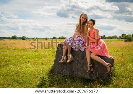 Two beautiful girls in the field. Girl sitting on a rock. They smile, radiate positive energy here, communicate. nature, beauty, girl, girlfriends - summer vacation concept. Article about the weekend.
