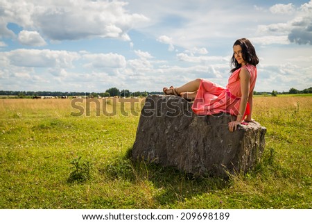 Beautiful girl in a red dress. Brunette sits on a rock, enjoying the outdoors and the sun. Country holiday, holiday,forest - The concept of country vacation. Article about the weekend in the country.