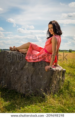 Beautiful girl in a red dress. Brunette sits on a rock, enjoying the outdoors and the sun. Country holiday, holiday,forest - The concept of country vacation. Article about the weekend in the country.