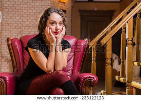 Portrait of beautiful romantic brunette on a red chair. Professional make-up on a beautiful woman. A girl sits in a beautiful  red chair and looks enigmatically. Romantic, beautiful girl portrait.
