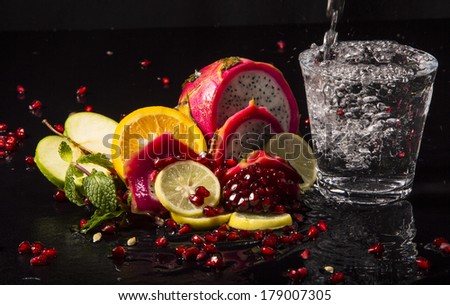 Colorful juicy assortment of sliced tropical fruits (purple dragon fruit, orange, lemon, pomegranate seeds, green apple and mint), on a wet black table, with a glass of transparent water