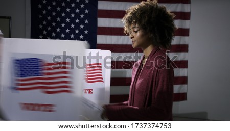 MS Young mixed-race woman ready to vote with blurred voting booth in foreground, being removed. Symbol of disenfranchisement with US flag backdrop