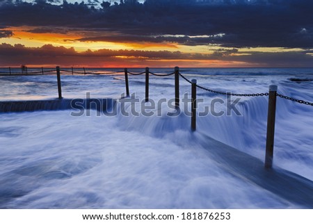 Ocean rock pool at sunrise with intense tidal wave overflowing above pool's edge with dramatic cloudy sky in Australia, Mona Vale beach