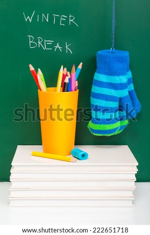 White books, colorful used pens in yellow glass and winter gloves on white desk against green chalkboard