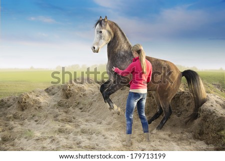Woman in red jacket and rising arabian horse on background of sand and fields