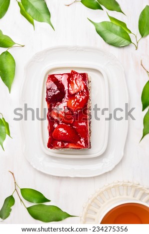 Piece of cake with strawberries and jelly, cup of tea and fresh green leaves on white wooden background