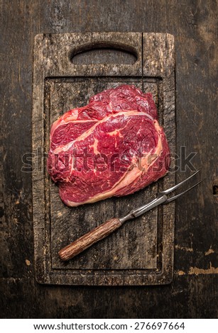 Two raw beef ribeye steak with meat fork on dark rustic wooden gutting board, top view