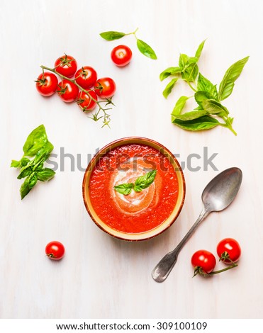 Bowl of tomato soup or gazpacho with spoon and basil on white wooden background, top view