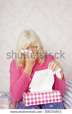 Sad woman with pink pajama and tissues sitting on bed blowing her nose