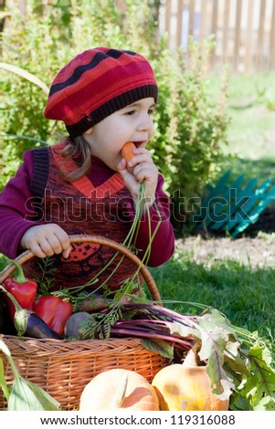 little girl sits on a grass near to a basket with vegetables and eats carrots