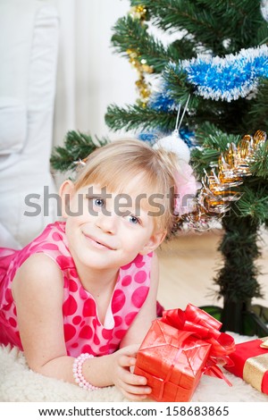 Red gift in hands of the fair-haired girl which sits near to a fur-tree