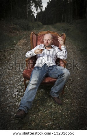 A thoughtful man / guy drinking cognac and smoke cigar in a vintage chair in the middle of a road in woods.