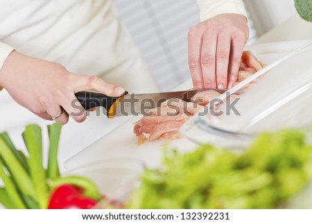 Young woman in a white kitchen chopping chicken meat fillet. Vegetable on the bench.