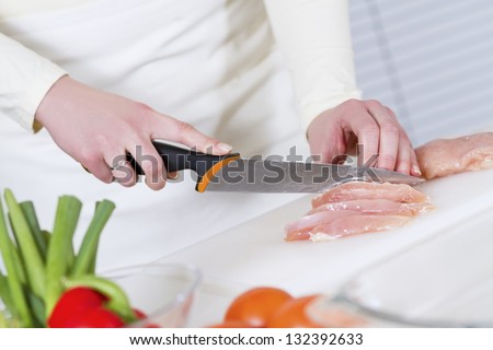 Young woman in a white kitchen cut chicken meat fillet. Vegetable on the bench.