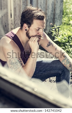 A redneck sitting outside a abandoned house and smoke cigarettes.