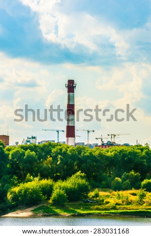 Chimney of a factory and construction site with cranes behind green trees. Industrial landscape with clouds in the sky and greenery.