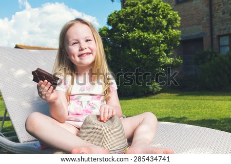 Little girl eaten chocolate on a sun lounger in the garden, in a sunny hot day.