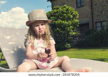 Little girl eaten chocolate on a sun lounger in the garden, in a sunny hot day.