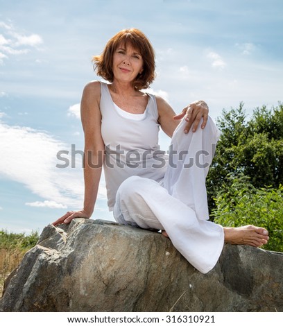 senior zen - smiling, beautifully aging woman sitting on a stone for outdoors yoga session wearing white seeking serenity and wellness in a park,summer daylight